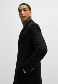 Cappotto in lana nera con design sartoriale, caratterizzato da un colletto alto e dettagli con bottoni. Maglione grigio a collo alto visibile sotto.