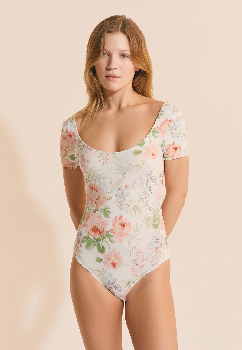 Frau mit hellbraunem Haar, die einen kurzärmeligen Bodysuit mit floralem Muster in Rosa und Grün vor beigem Hintergrund trägt.