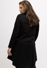 Tunica nera a maniche lunghe con orlo arrotondato e texture liscia, che mostra un design semplice e minimalista che arriva fino a metà coscia.