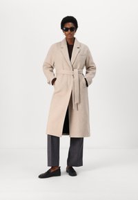 Cappotto beige con cintura realizzato in tessuto texturizzato, con colletto a rever e tasche laterali, abbinato a pantaloni antracite e scarpe nere.
