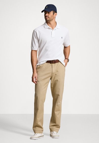 T-shirt polo blanc, pantalon beige légèrement usé, ceinture marron et baskets blanches. Casquette marine avec logo, pose debout contre un fond clair.