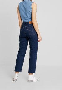 Jeans en denim bleu foncé taille haute avec une coupe droite, poches arrière discrètes et un patch en cuir sur la taille. Paire de baskets blanches.