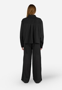 Camicia nera ampia con schiena plissettata e polsini con bottoni, abbinata a pantaloni neri a gamba larga. Tessuto liscio in tutta la superficie.
