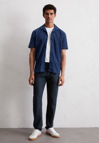 Man die tegen een witte muur staat, gekleed in een marineblauwe short-sleeve overhemd, een witte t-shirt, donkere broek en witte sneakers met tan zolen.