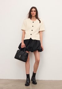 Woman wearing short-sleeve cream buttoned jacket, black puffed skirt, black ankle boots, holding black leather handbag against plain wall background.