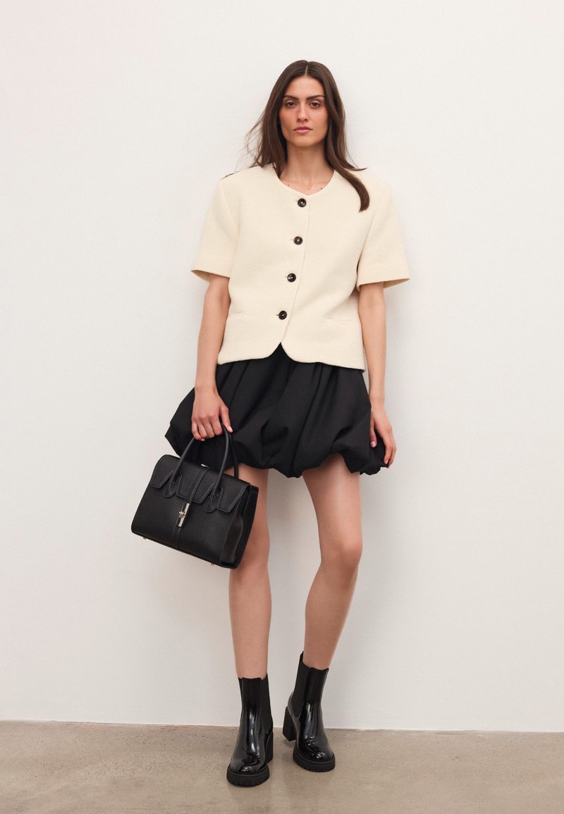 Woman wearing short-sleeve cream buttoned jacket, black puffed skirt, black ankle boots, holding black leather handbag against plain wall background.