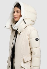 Daunenjacke in hellem Creme mit einem flauschig weiß gefütterten Kapuzen. Verfügt über einen Reißverschluss vorn, Druckknöpfe und ein Logo-Patch am Ärmel.