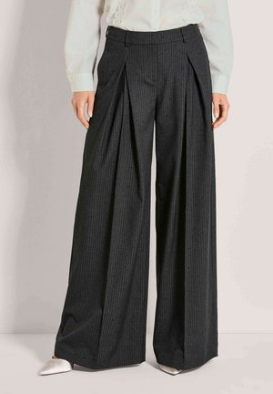 Frau trägt hochtaillierte, weit geschnittene schwarze Nadelstreifenhose mit Vorderfalten, kombiniert mit weißen spitzen Schuhen und einer weißen Bluse.