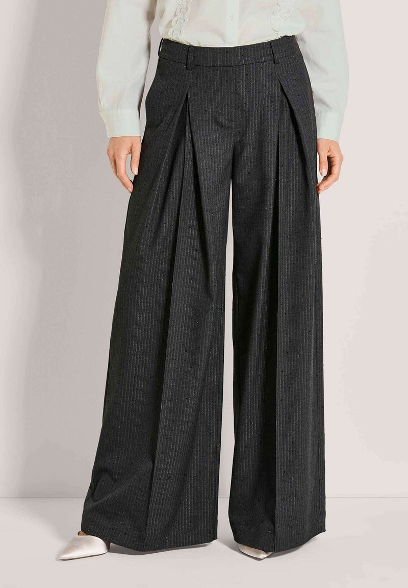 Frau trägt hochtaillierte, weit geschnittene schwarze Nadelstreifenhose mit Vorderfalten, kombiniert mit weißen spitzen Schuhen und einer weißen Bluse.