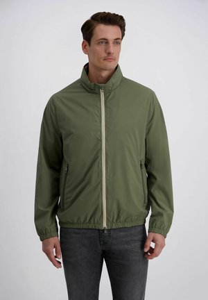 Man draagt een olijfgroene jas met ritssluiting, ritssluiting-zijzakken en elastische manchetten, gecombineerd met donkergrijze jeans, staand tegen een witte achtergrond.