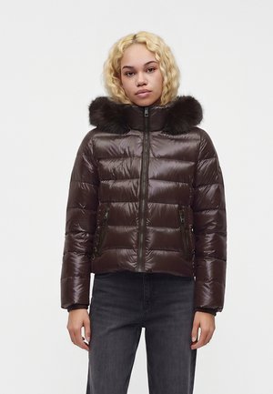 Junge Frau mit blondem lockigem Haar trägt eine dunkelbraune gesteppte Jacke mit pelzbesetzter Kapuze und schwarze Jeans vor einem schlichten weißen Hintergrund.