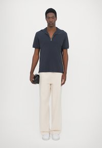 Navy short-sleeve zip polo, cream wide-leg trousers, and white sneakers. Model holds a black textured clutch. Simple, modern design.