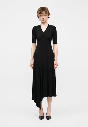 Νεαρό άτομο που στέκεται, φορώντας μαύρο ασύμμετρο midi φόρεμα με μανίκια μέχρι τον αγκώνα και μαύρες μυτερές γόβες, σε λευκό φόντο.