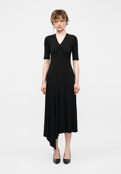 Jeune personne debout, portant une robe midi noire asymétrique avec des manches jusqu'au coude et des talons pointus noirs sur fond blanc.