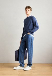 Sudadera azul marino, camiseta interior azul clara, jeans sueltos azules, zapatillas blancas, llevando un bolso oscuro con correas marrones, con un fondo neutro.