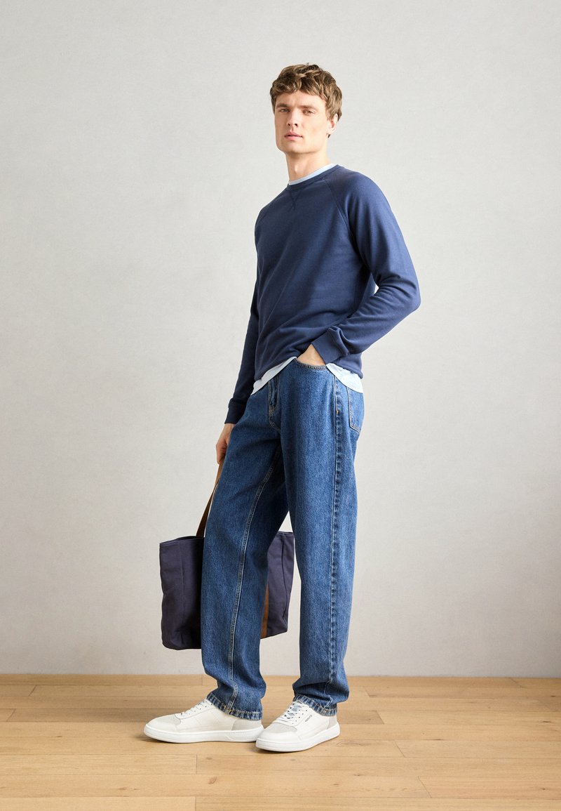 Felpa blu marina, maglietta azzurra sotto, jeans blu larghi, sneakers bianche, in mano una borsa scura con cinturini marroni, su uno sfondo neutro.