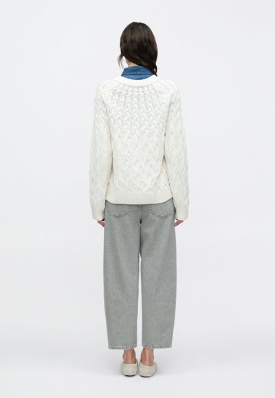 Πλεκτό πουλόβερ σε κρεμ απόχρωση με σχέδιο κοτσίδας και ριμπ τελείωμα, συνδυασμένο με ανοιχτό γκρι τζιν παντελόνι με φαρδύ πόδι. Φαίνεται από πίσω, με ορατό μπλε γιακά.