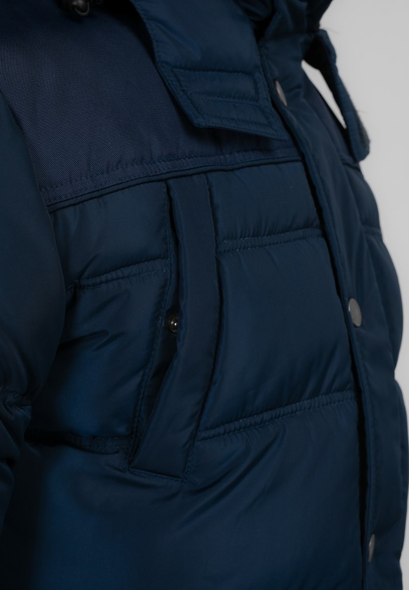 Veste rembourrée bleu marine avec un design matelassé, présentant un mélange de tissus lisses et texturés, des poches à pression et un col ajustable.