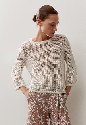 Frau mit Dutt trägt einen weißen, offen gestrickten Pullover und braune, gemusterte weite Hosen, Hände in den Taschen, schaut seitlich nach unten.