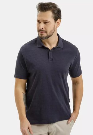 Mężczyzna z brodą, ubrany w granatową koszulkę polo z krótkim rękawem i beżowe spodnie, stoi z jedną ręką w kieszeni, patrząc na bok.