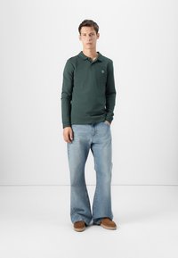 Musta vihreä pitkähihainen polo-paita kauluksella, yhdistettynä vaaleansinisiin leveälahkeisiin farkkuihin ja ruskeisiin liukuhihnasandaaleihin. Yksinkertaiset kankaan tekstuurit.