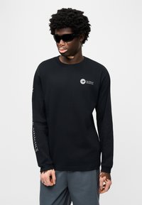 Μαύρο T-shirt με μακριά μανίκια από βαμβάκι, με μικρό λογότυπο στο στήθος και κείμενο κατά μήκος του αριστερού μανικιού. Φορώντας γυαλιά ηλίου και γκρι παντελόνι.