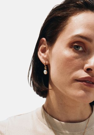 Kullast rõngakujulised kõrvarõngad, millel on keerlev muster ja üks rippuv pärl. Modelli juuksed on lühikesed ja ta kannab kerget kampsunit.