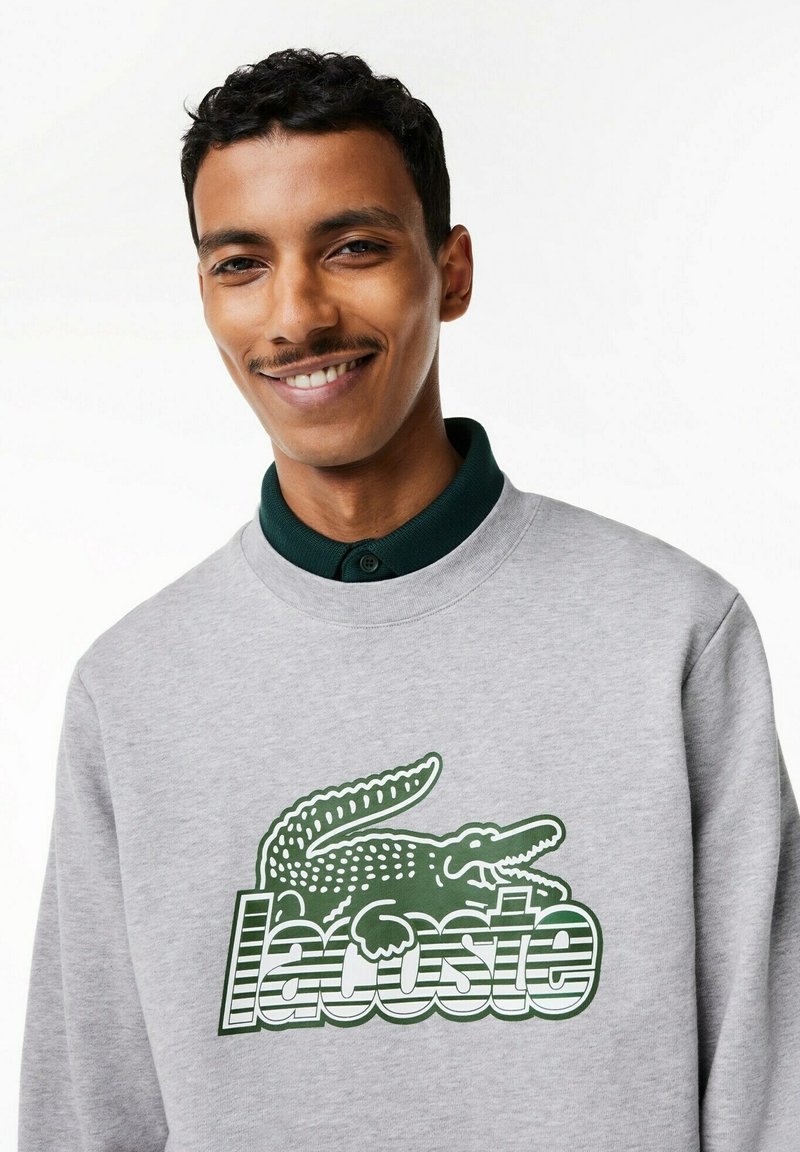 Felpa grigia con un logo del coccodrillo verde in evidenza e il testo "Lacoste", realizzata in un tessuto testurizzato e con colletto a costine di colore verde scuro.