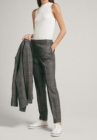 Femme portant un pantalon à carreaux gris et un top sans manches blanc, tenant une veste à carreaux assortie d'une main, portant des baskets blanches.