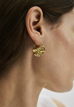 Prim-plan al urechii unei femei purtând o cercei texturată, aurie, în formă de semilună, cu finisaj ciocanit, pe piele deschisă la culoare și cu păr castaniu.