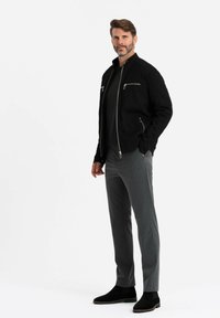 Veste zippée noire avec un col montant, associée à un pantalon gris sur mesure et des bottines en daim noir. Design simple, sans motifs.