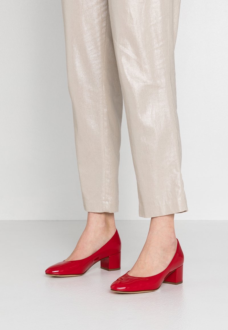 Chaussures à talons carrés en cuir verni rouge, bout arrondi avec découpe, associées à un pantalon beige clair, brillant et raccourci.