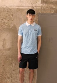 Hellblaues Poloshirt mit Rautenmuster, weißem Kragen und Logo; kombiniert mit schwarzen Shorts mit Taschen. Einfaches, lässiges Design.