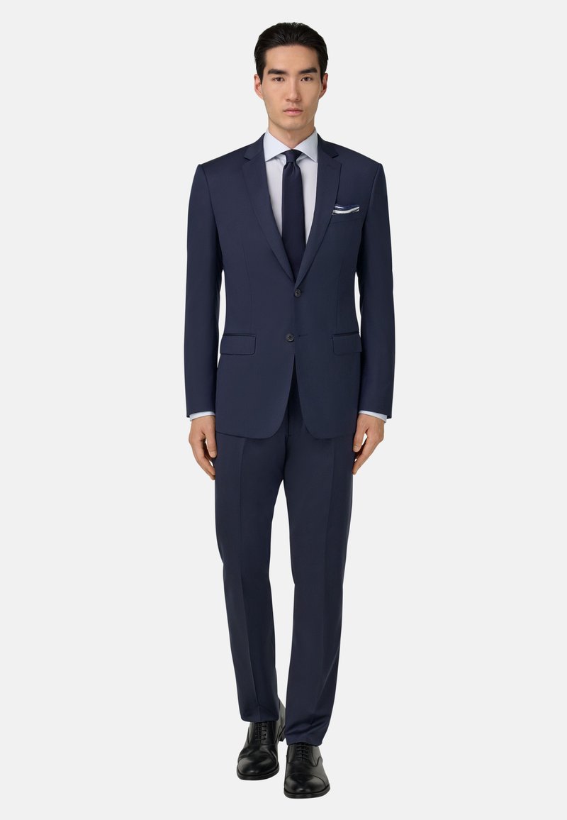 Tamsiai mėlynas kostiumas su įpjovos apykakle, dviejų mygtukų švarkas, plokščių priekių kelnės, derinamas su šviesiai mėlyna marškiniais ir tamsia kaklaraiščiu. Juodi batai.