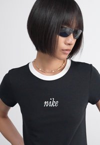 Fekete pamut póló fehér kerek nyakkal, rajta írásos "nike" logóval. A modellen fekete napszemüveg és aranylánc nyaklánc található.