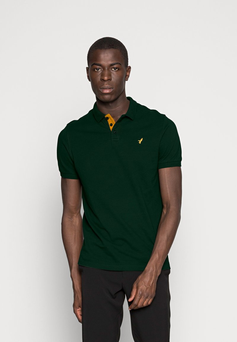 Dunkelgrünes Poloshirt mit einem gelben Knopfkragen und einem kleinen gestickten Logo auf der linken Brust. Kurze Ärmel und eine klassische Passform.
