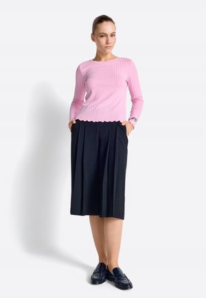 Kobieta ubrana w różowy, prążkowany top z długim rękawem, czarną plisowaną spódnicę do kolan i czarne mokasyny, stojąca z rękami w kieszeniach.