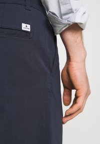 Mörkblå byxor med en slät yta, med en bakficka och en liten etikett. Hand som vilar bredvid midjan. Ljusblus skjorta synlig.