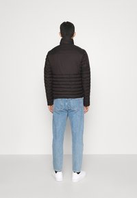 Černá péřová bunda s prošívanou texturou a vysokým límcem, v kombinaci se světle modrými džíny a bílými teniskami, pohled zezadu.