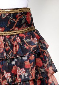 Bloemen tiered rok van doorschijnende stof met zwarte, rode en blauwe tinten en een gouden pailletten tailleband. Getextureerde ruches en ingewikkelde patronen.