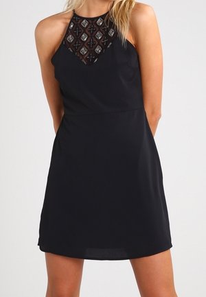 Robe noire à encolure haute, ornée d'une bordure embellie et à motifs. Tissu lisse avec une jupe évasée coupée au-dessus du genou.
