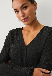 Schwarzes Kleid mit V-Ausschnitt, das mit schimmernden silbernen Punkten verziert ist. Der Stoff ist glatt und flexibel, mit langen, sanft gerafften Ärmeln.