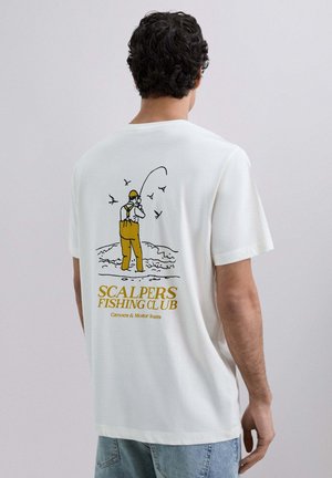 Hombre con cabello rizado y oscuro que lleva una camiseta blanca con una ilustración de pesca en amarillo y negro y el texto "Scalpers Fishing Club" en la parte de atrás.