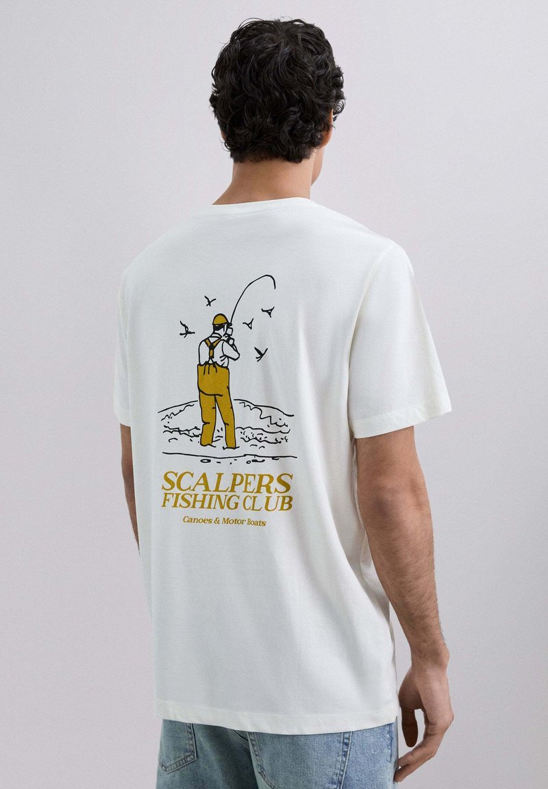 Bărbat cu păr creț închis la culoare, purtând un tricou alb cu o ilustrație de pescuit galbenă și neagră și textul "Scalpers Fishing Club" pe spate.
