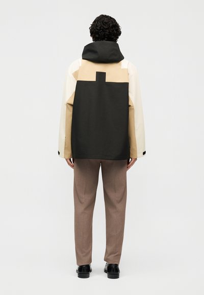 Άνδρας όρθιος με την πλάτη προς το φακό, φοράει ένα χρωματιστό μπουφάν σε μπεζ, μαύρο και άσπρο με κουκούλα, καφέ παντελόνι και μαύρα παπούτσια.