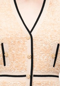 Ροδακινί πλεκτή ζακέτα με μαύρες και μπεζ λεπτομέρειες, διακοσμημένη με χρυσά κουμπιά και δύο μπροστινές τσέπες σε έναν κορμό ανθρώπου.