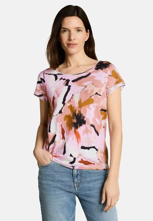 Woman wearing a pink, orange, white, and black abstract patterned short-sleeve top with blue jeans, standing with one hand in pocket.