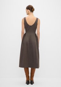 Robe marron longueur genou avec un dos en V, sans manches, côtés plissés et texture lisse, assortie à des bottes marron jusqu'au genou.