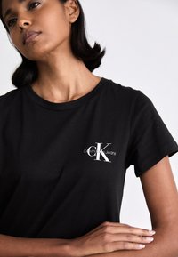 Black cotton t-shirt with a crew neckline, featuring a white "Calvin Klein Jeans" logo on the left chest area. Finely textured fabric.