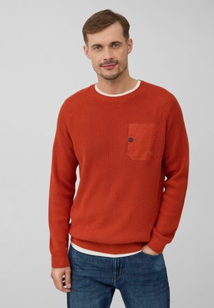 Roter strukturierten Strickpullover mit rundem Ausschnitt, der eine Brusttasche und einen kontrastierenden hellen Innenkragen aufweist. Kombiniert mit blauen Jeans.